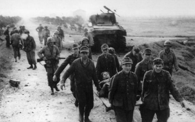 Prisioneros alemanes transportando un herido. Al fondo un Sherman puesto fuera de combate